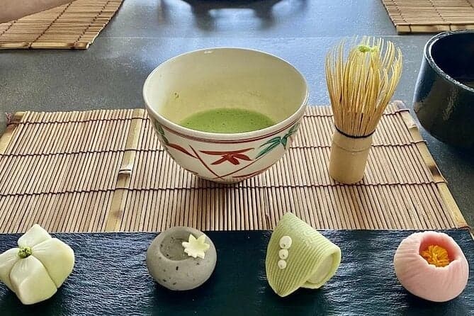 A bowl of matcha tea with a tea whisk, reminiscent of a Japanese tea ceremony, is accompanied by four pieces of wagashi on a mat-covered table.