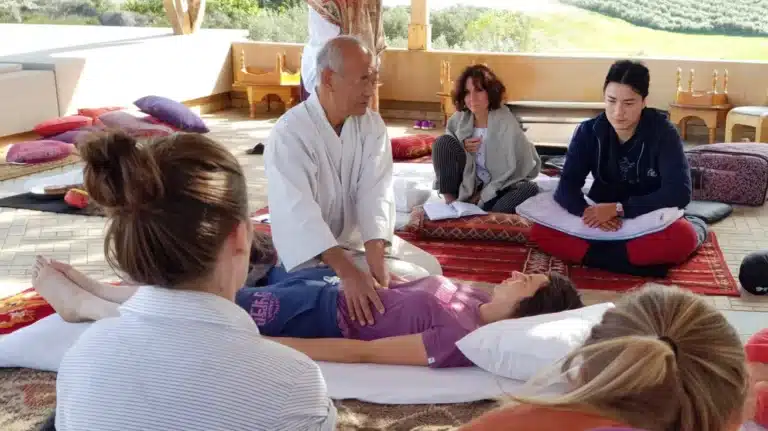 An older man in white clothes performs a healing or massage technique on a woman lying on a mat, while others observe and take notes in this shiatsu massage school group setting.