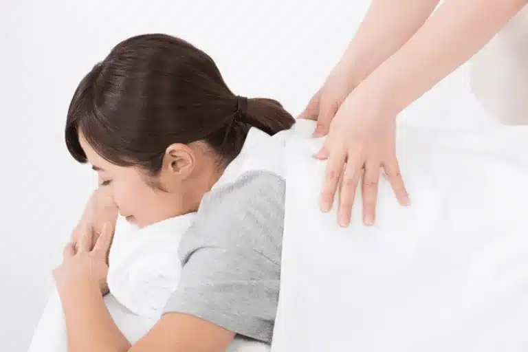 Person lying face down receiving a relaxing back massage from another person, partially covered with a white towel.