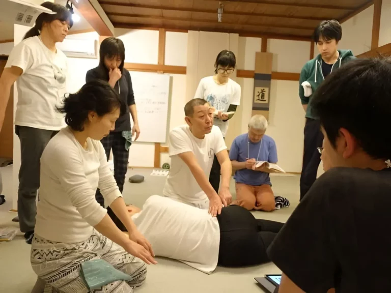 A group of people in a room observe as an instructor at a shiatsu massage school demonstrates a bodywork technique on a person lying face down.