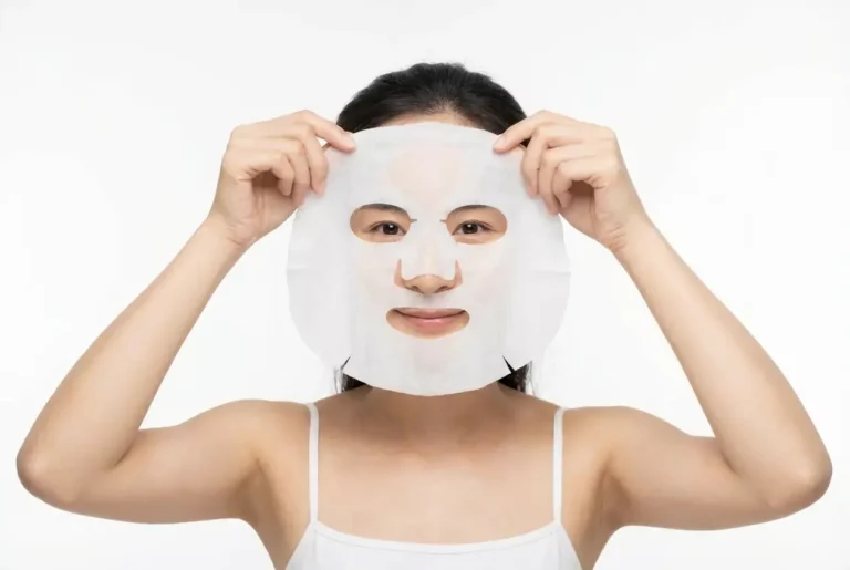 A person wearing a white vest is applying a sheet mask to their face against a plain white background, embracing a relaxing skincare routine inspired by onsen Singapore.