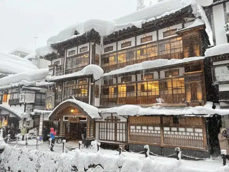 A traditional Japanese building with warmly lit windows, reminiscent of an onsen, is covered in heavy snow as several people walk outside in the snowy weather.