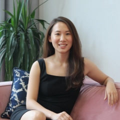 A woman with long brown hair in a black dress sits on a pink couch, smiling, with a blue and white pillow and a green plant in the background.