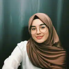 A woman wearing glasses and a brown hijab over a white top smiles slightly while sitting in front of a dark background.