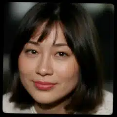 A woman with straight dark hair and bangs, wearing a light-colored top, looks at the camera with a neutral expression.