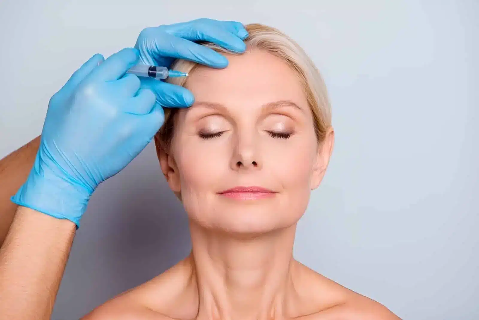 A woman with closed eyes receives a botox injection in her forehead from a person wearing blue gloves, against a plain light background.