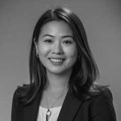 A woman with medium-length hair, wearing a blazer and necklace, smiles at the camera against a plain background. The image is in black and white.