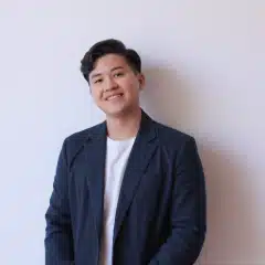 Person with short dark hair, wearing a navy blazer over a white shirt, standing against a plain light-colored wall and smiling at the camera.