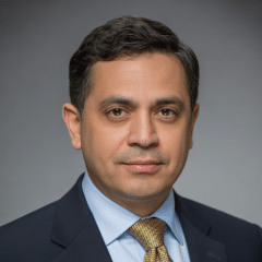 A man in a suit and tie poses for a professional headshot against a plain gray background.