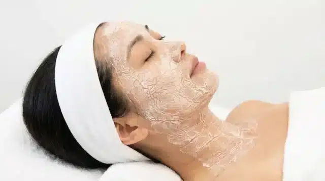 A woman lies with eyes closed, wearing a white headband and dressing gown, while a clear drying lifting facial mask covers her face and neck.