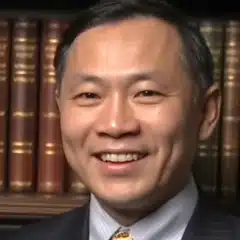 A man in a suit smiles at the camera, with a background of books on a shelf.