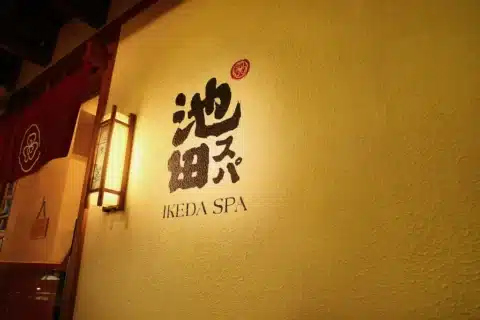 Wall with Japanese characters and the English text "IKEDA SPA" next to a lantern-style light fixture, reflecting the spirit of omotenashi in an inviting indoor setting.