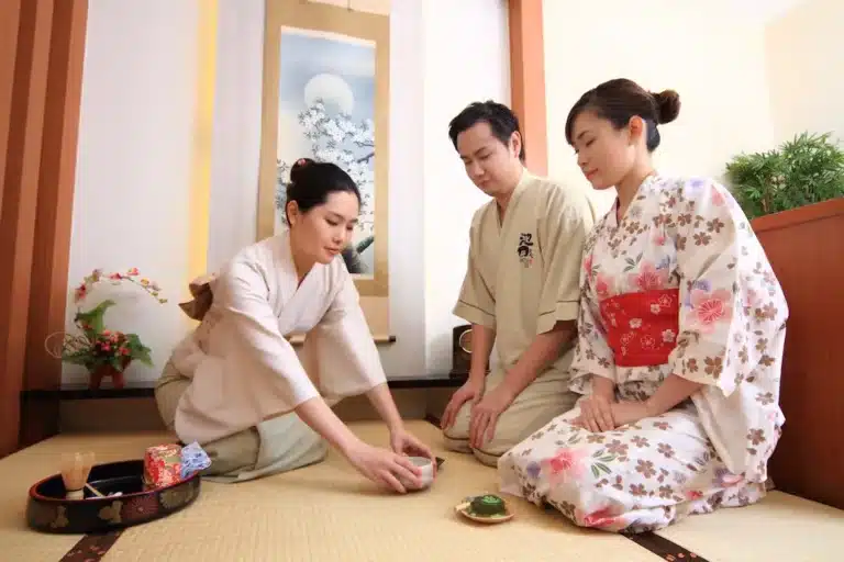 Three people in traditional Japanese attire embody omotenashi as they participate in a tea ceremony, sitting on a tatami mat with tea utensils arranged before them.