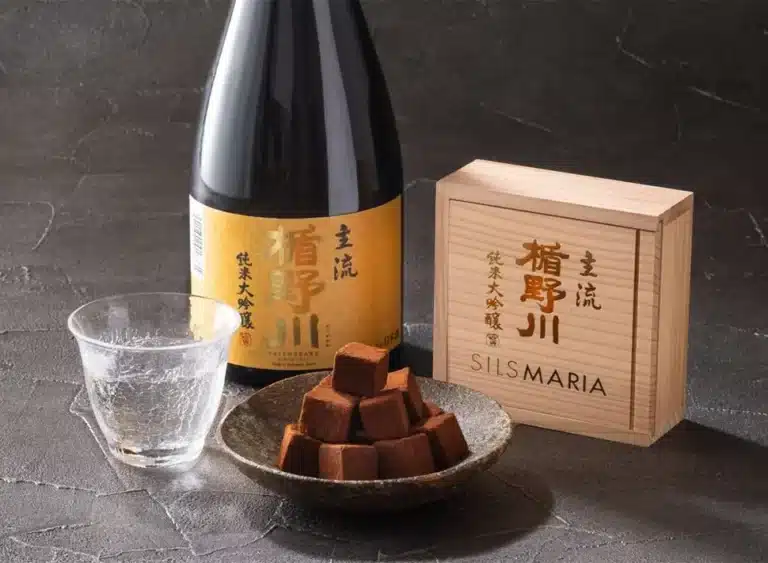 A bottle of sake, a glass, a wooden box, and a plate of chocolate truffles are arranged on a dark surface.