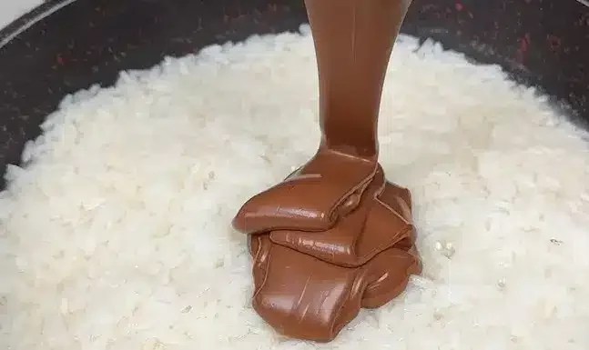 Chocolate being poured onto a bed of uncooked white rice in a dark-coloured pan.