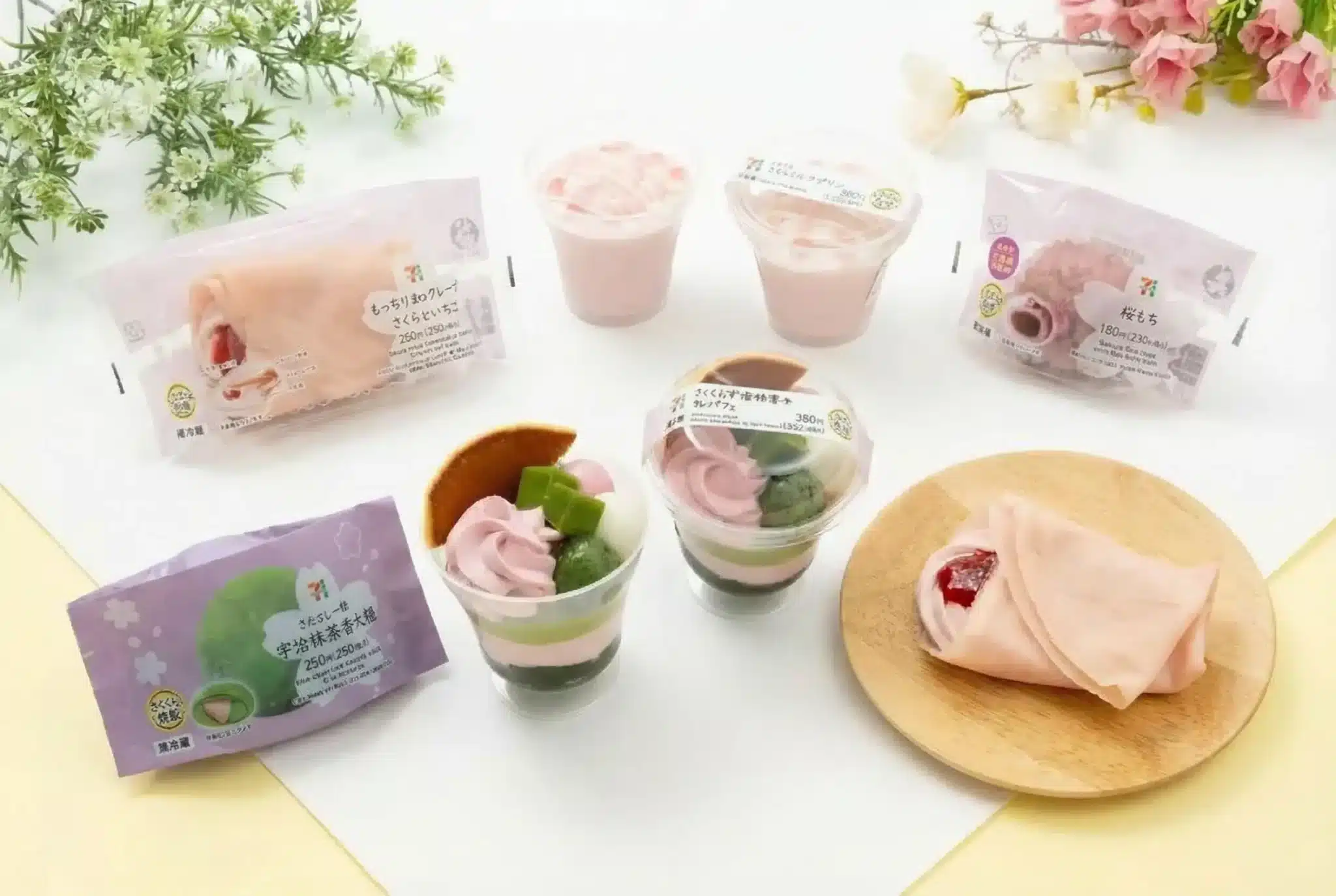 A selection of pink and green Japanese-style desserts, inspired by the cherry blossom forecast, including cups, packaged treats, and crêpes, is arranged on a light-coloured surface with flowers as decoration.