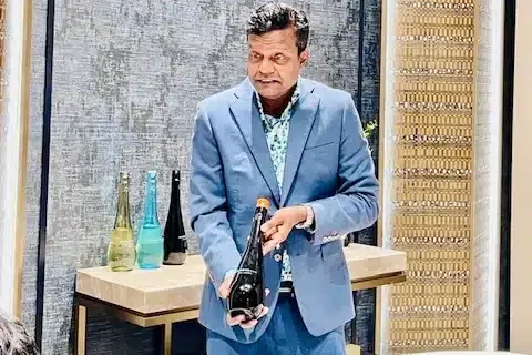 A man in a blue suit holds a dark bottle, standing in front of a table with several other bottles in a modern room.