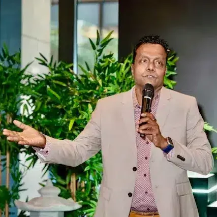 A man in a beige suit speaks into a microphone while gesturing with his hand, standing in front of green indoor plants.