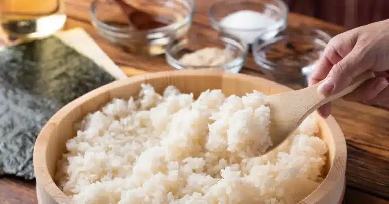 A hand uses a wooden spoon to scoop cooked white rice from a wooden bowl, with nori sheets and small bowls of ingredients in the background.
