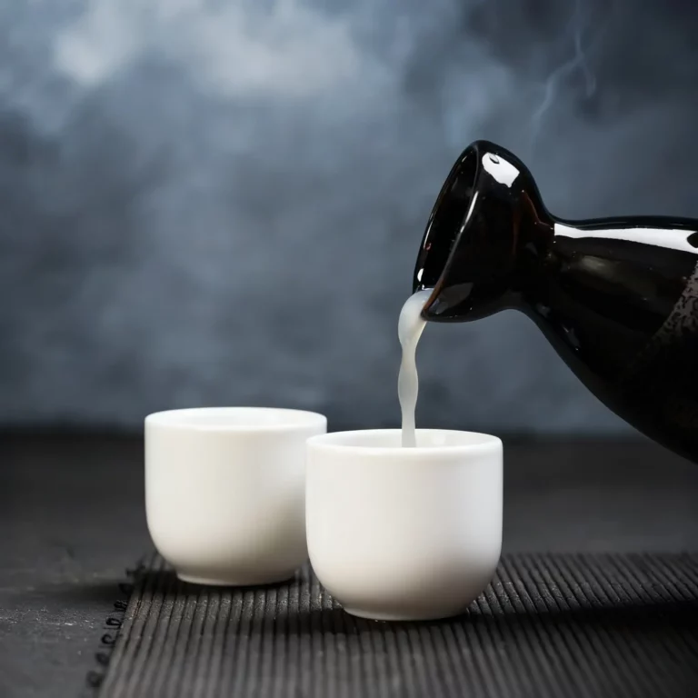 A black sake bottle pours steaming sake into one of two white cups on a dark surface with a textured placemat, perfect for a relaxing sake chocolate pairing.
