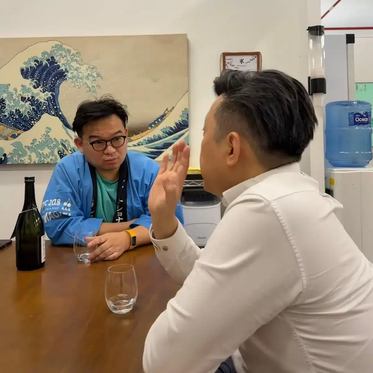 Two men sit at a table having a conversation, with a bottle and two glasses—perhaps discussing sake chocolate pairing. A painting of a wave and a water dispenser are visible in the background.