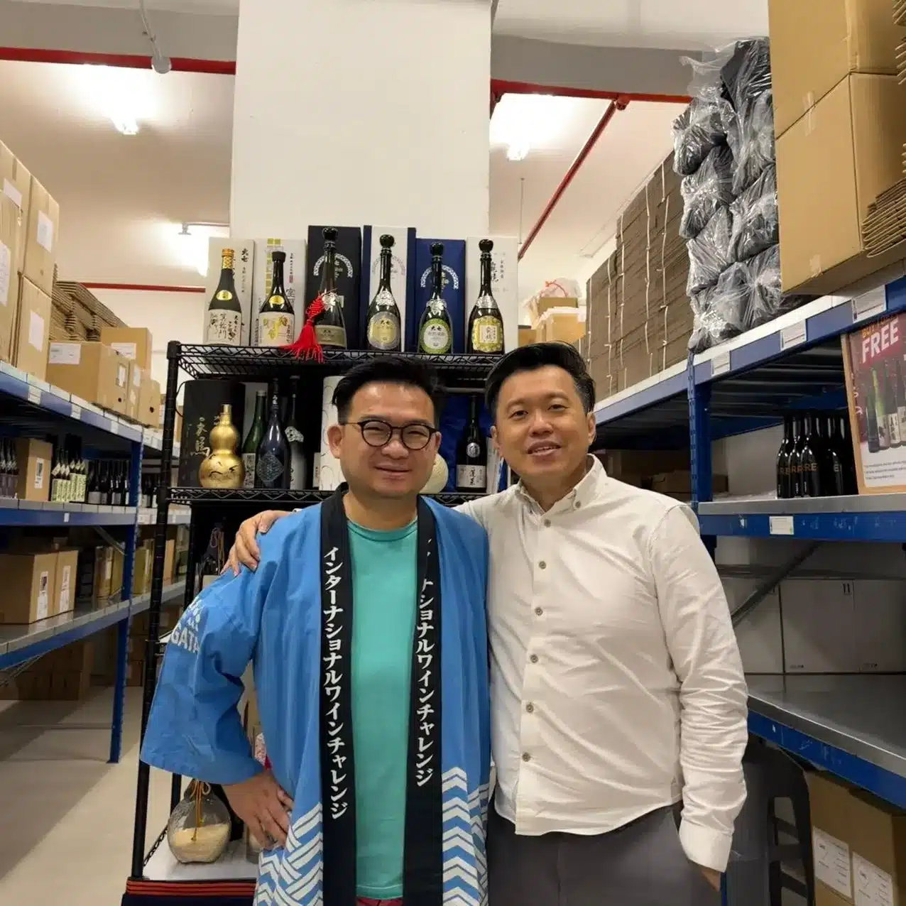 Two men stand smiling in a storeroom with shelves holding bottles and boxes, perhaps preparing for a sake chocolate pairing or savouring the latest sake Kit Kat release. One wears a patterned dressing gown, the other a white shirt.