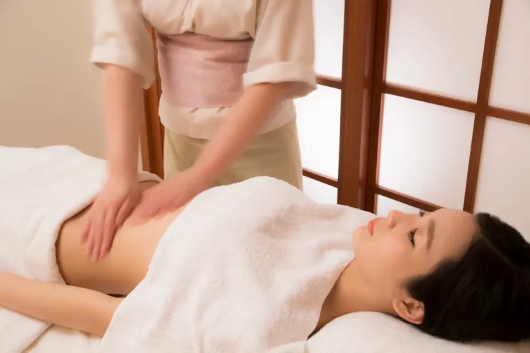 A person lying on a massage table covered with a towel receives an abdominal massage from a skilled massage therapist in a serene spa setting.