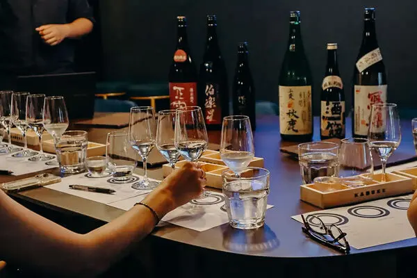 A table set for a sake tasting event, featuring several bottles of sake, rows of glasses, water glasses, tasting mats, and a special sake chocolate pairing experience, with people seated round the table.
