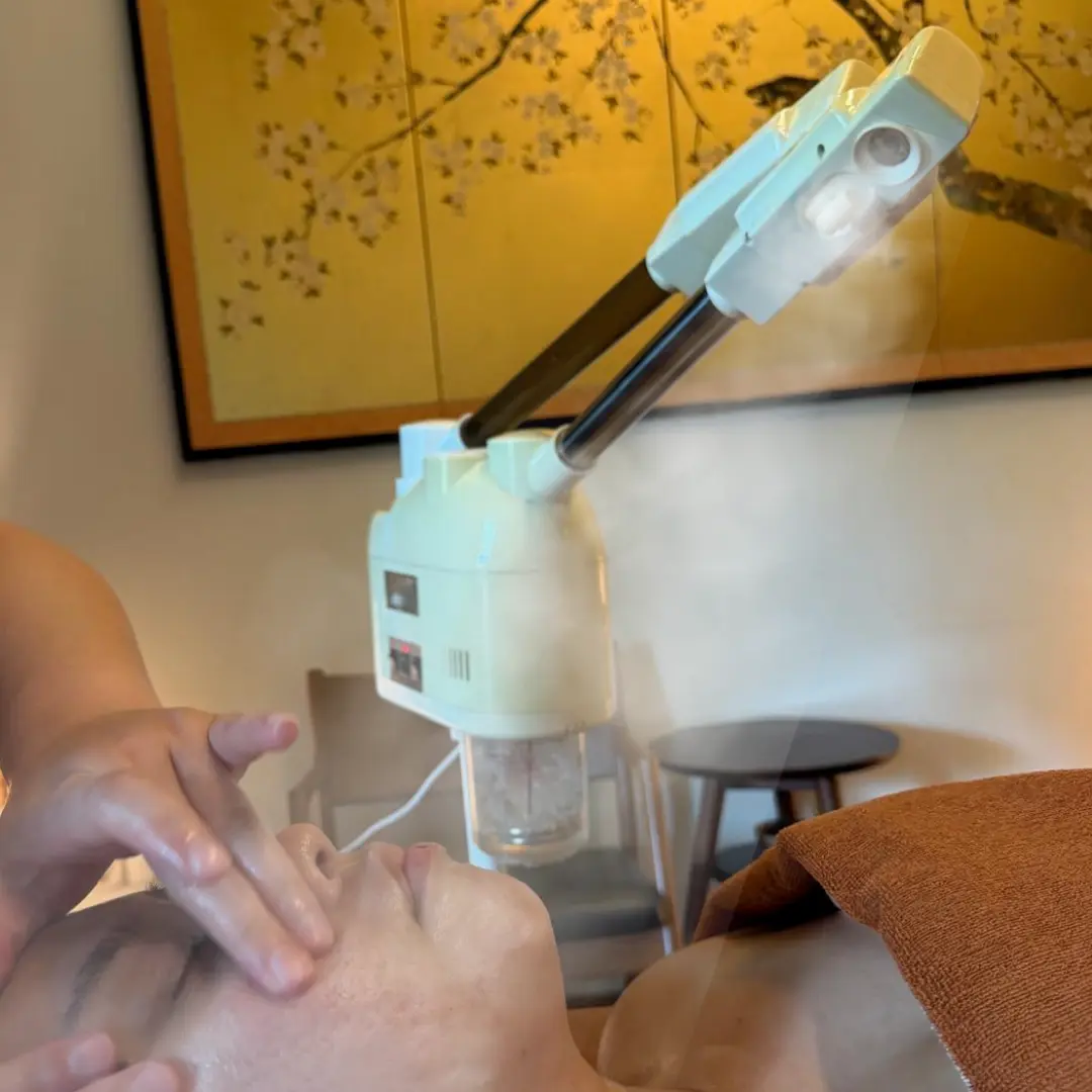 A person receives a deep cleansing acne facial with steam applied to their face, whilst a hand gently massages their cheek. A decorative painting hangs on the wall in the background.