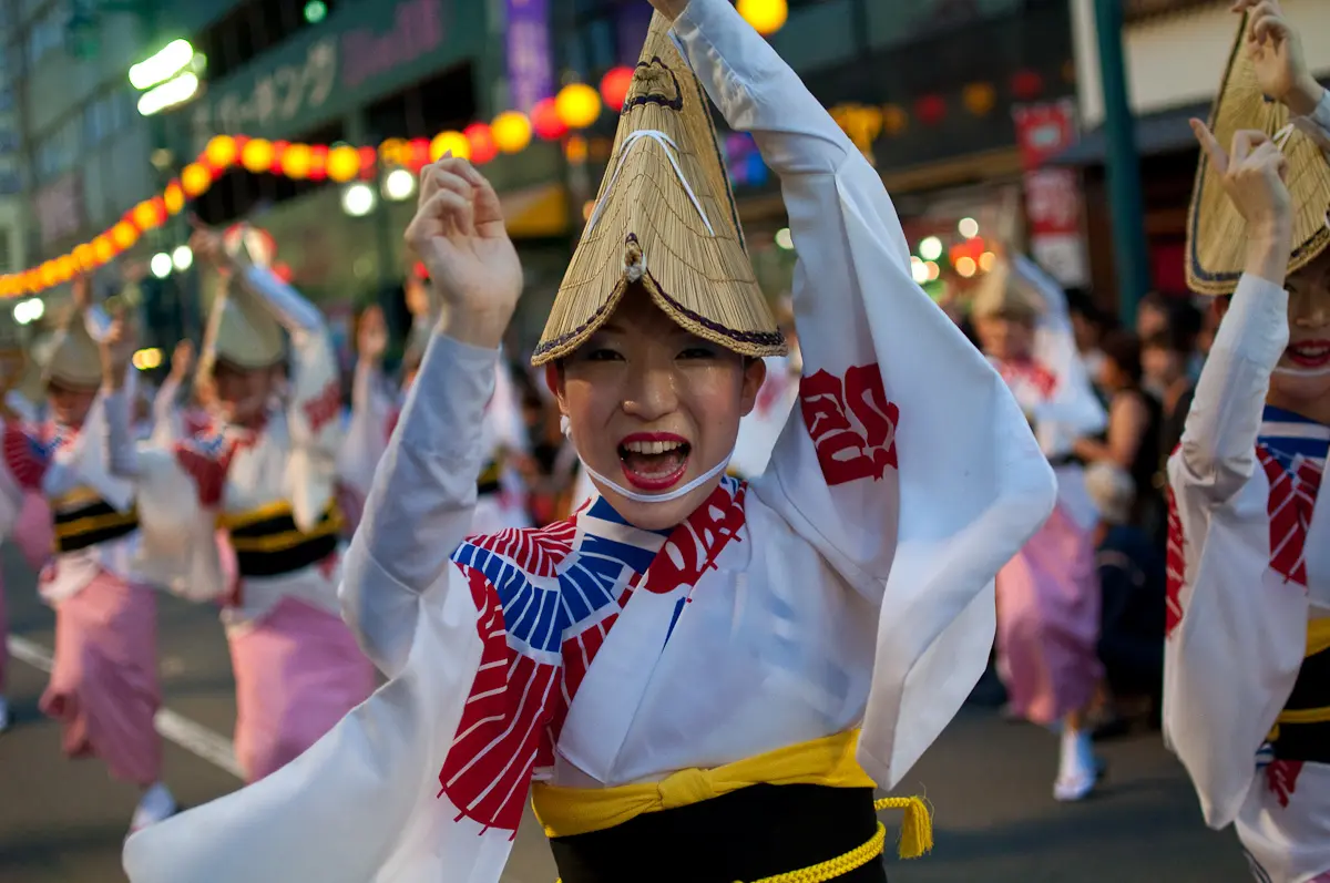 A group of dancers in traditional Japanese attire and straw hats perform at a street festival, with colourful lanterns, ramune soda bottles, and spectators in the background.