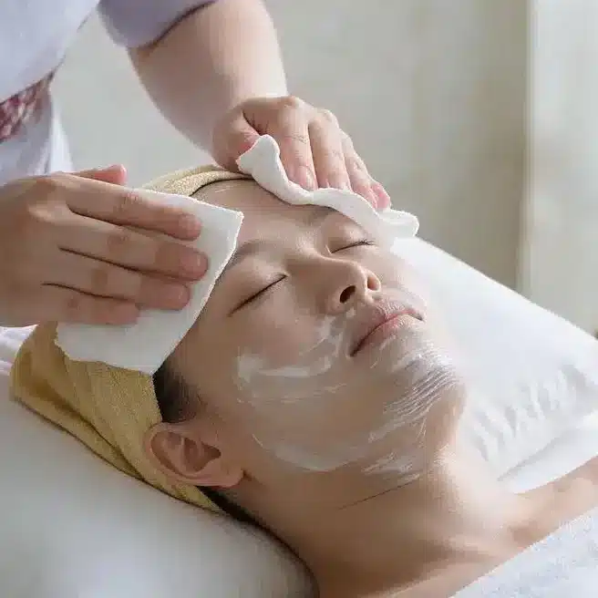 A person lying on a spa bed receives an onsen facial cleanse from a staff member using cotton wool pads; skincare products and towels are nearby. Text reads: “Step 1: Double Cleanse.”.