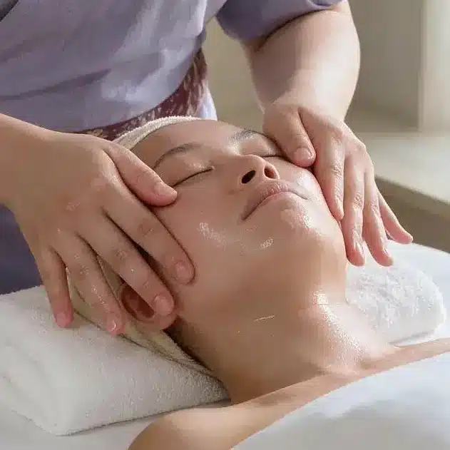 A person receives an onsen facial massage on a spa bed, with a staff member applying oil and pressing key points on the face; skincare products are visible nearby.