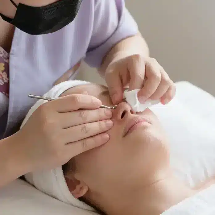 A person wearing a mask and purple uniform performs an onsen facial treatment on a client lying on a massage table with a white towel and headband.
