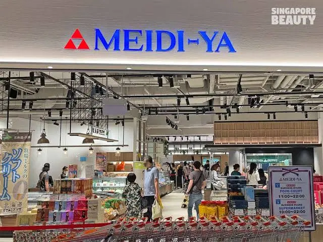 People shopping inside a brightly lit MEIDI-YA supermarket, with shopping trolleys and food products—including colourful bottles of ramune—visible near the entrance.