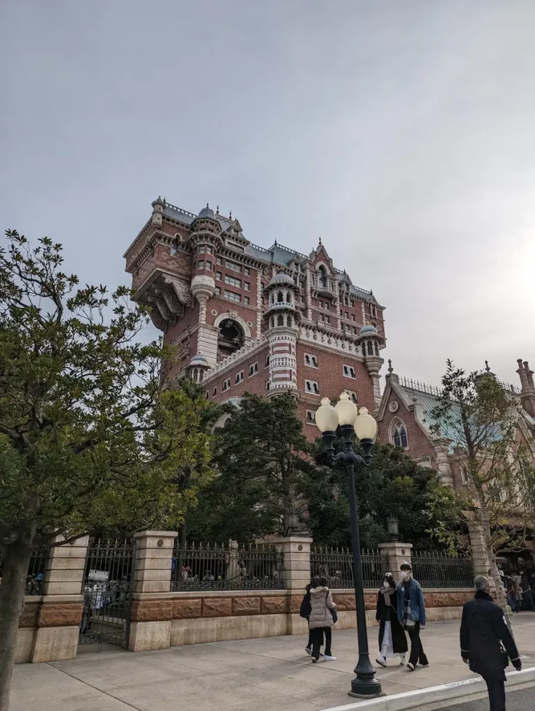 A tall, ornate brick building with a unique extended upper section reminiscent of the enchanting architecture found at DisneySea, surrounded by trees and people walking on the pavement below.