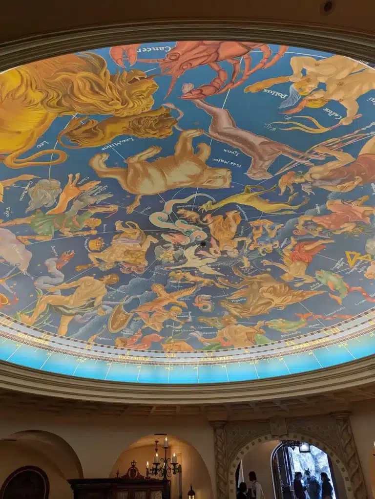 Circular ceiling mural depicting colourful zodiac constellations on a blue background, reminiscent of the enchanting atmospheres found at DisneySea, viewed from below in a grand ornate room with an arched entryway and visitors.