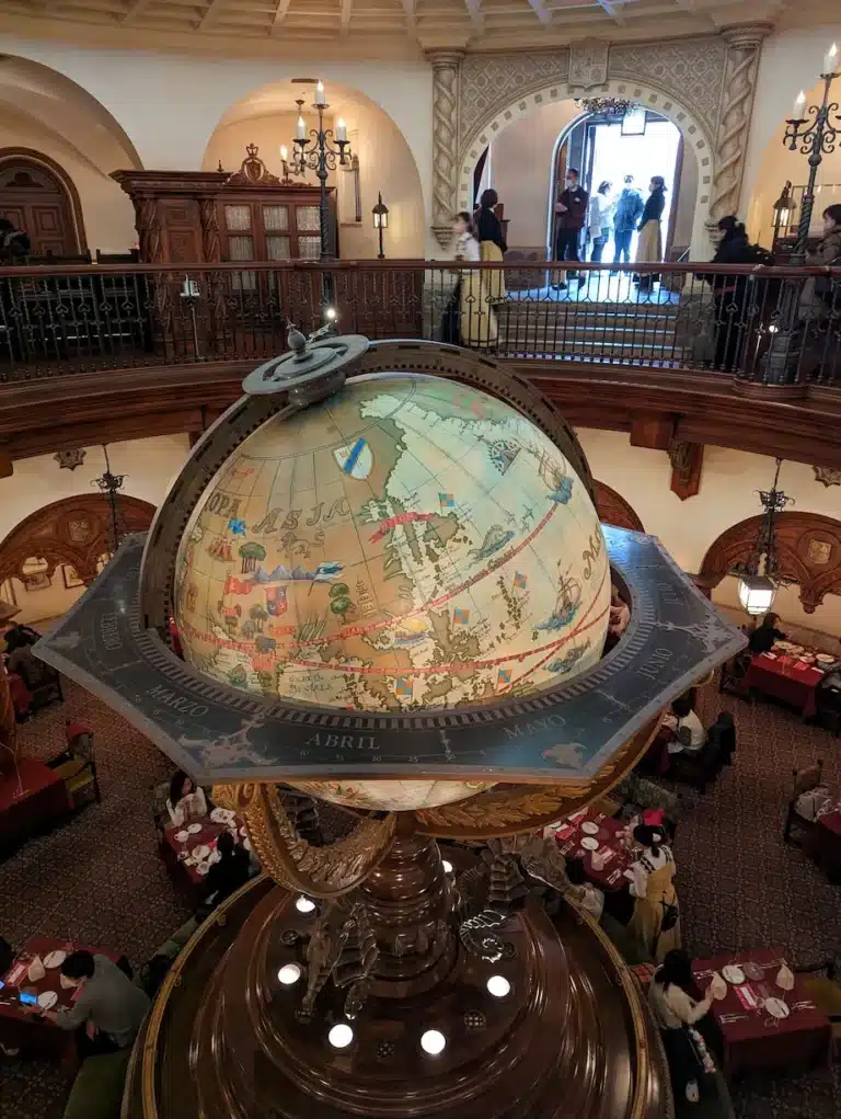 Large vintage globe with zodiac ring centrepiece in a grand, ornate restaurant at DisneySea; diners are seated below and people are visible on an upper balcony near a bright entrance.