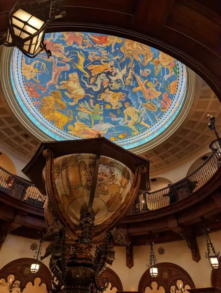 A large, ornate globe stands beneath a domed ceiling featuring a colourful zodiac mural, reminiscent of the enchanting atmosphere found at DisneySea, surrounded by a wooden balcony and decorative lighting.