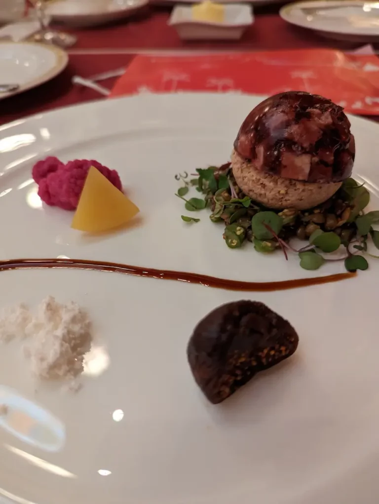 A plated gourmet appetiser inspired by DisneySea, featuring a small dome, pink purée, yellow cube, dark fig, white powder, microgreens, and a brown sauce line artfully arranged on a white dish.