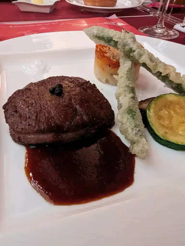 A plated steak with brown sauce, tempura green beans, a round potato stack, and a slice of courgette is artfully arranged on a white square plate—reminiscent of the beautifully presented dishes found at DisneySea.