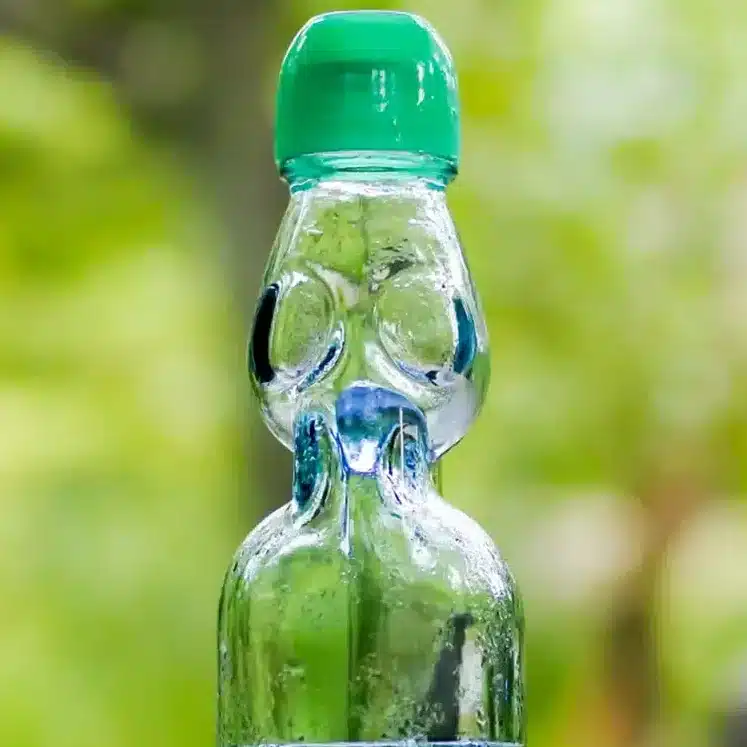 A clear glass ramune bottle with a green cap, partially filled with liquid, sits on an outdoor wooden table with a blurred green background.