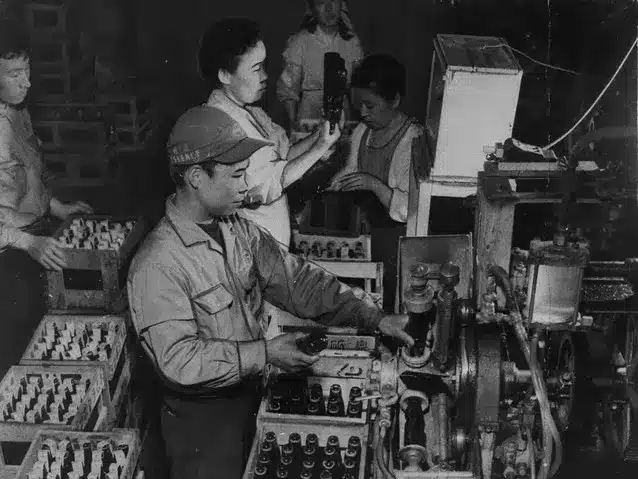 Workers operate machinery and handle glass bottles of ramune on an assembly line in a bottling factory, surrounded by crates filled with bottles.