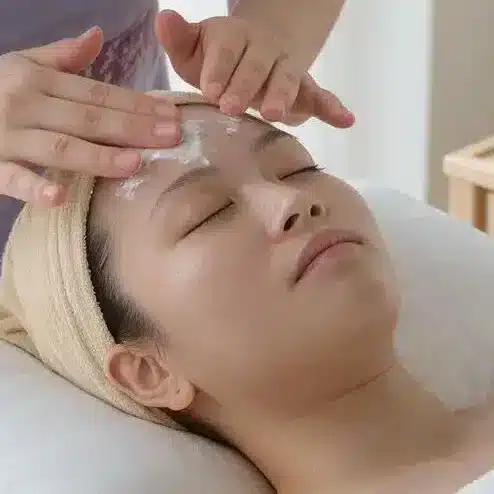 A person in a spa receives an onsen facial sunscreen application on their face from a staff member wearing a purple uniform; skincare products and towels are on a nearby table.