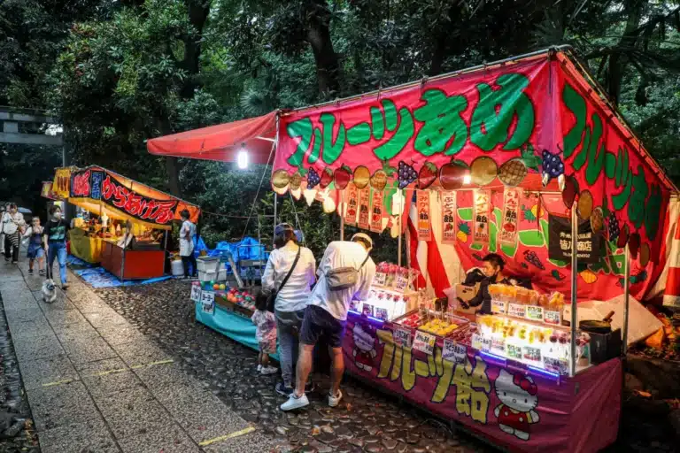 Japanese outdoor market stalls with colourful signs and hanging lanterns, selling snacks, sweets, and refreshing bottles of ramune, surrounded by trees as people browse and make purchases on a stone pathway.