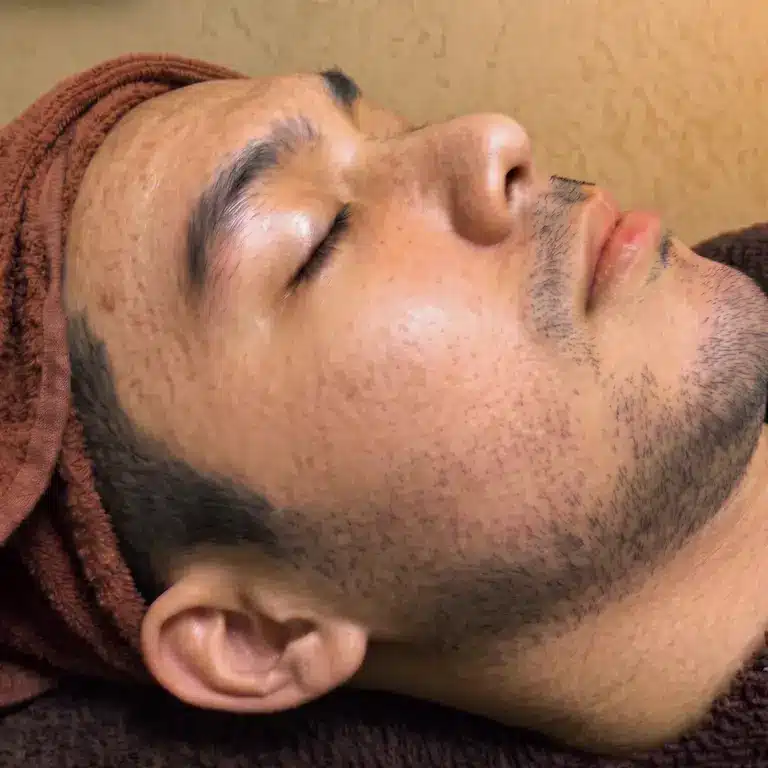 A man with a towel wrapped around his head lies with his eyes closed, appearing relaxed against a brown background, as if enjoying the soothing effects of an acne facial treatment.