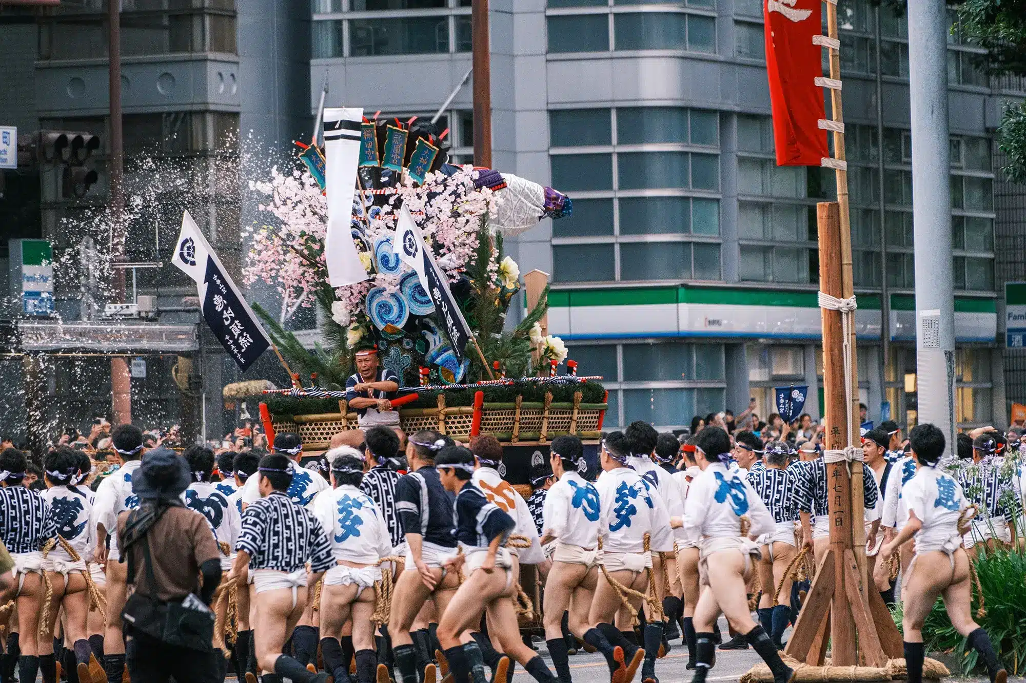 A group of people in traditional attire carry a decorated float during a festival parade on a city street, with water splashing nearby and bottles of ramune clinking cheerfully in their hands.
