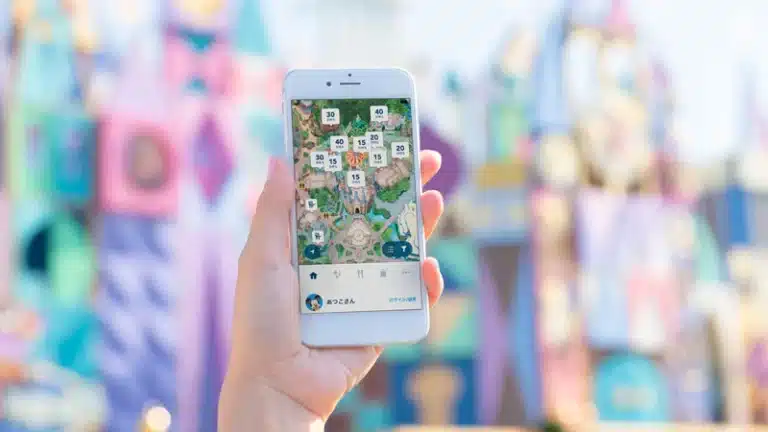 A hand holds a smartphone displaying a DisneySea theme park map with queue times for various attractions; colourful buildings are blurred in the background.