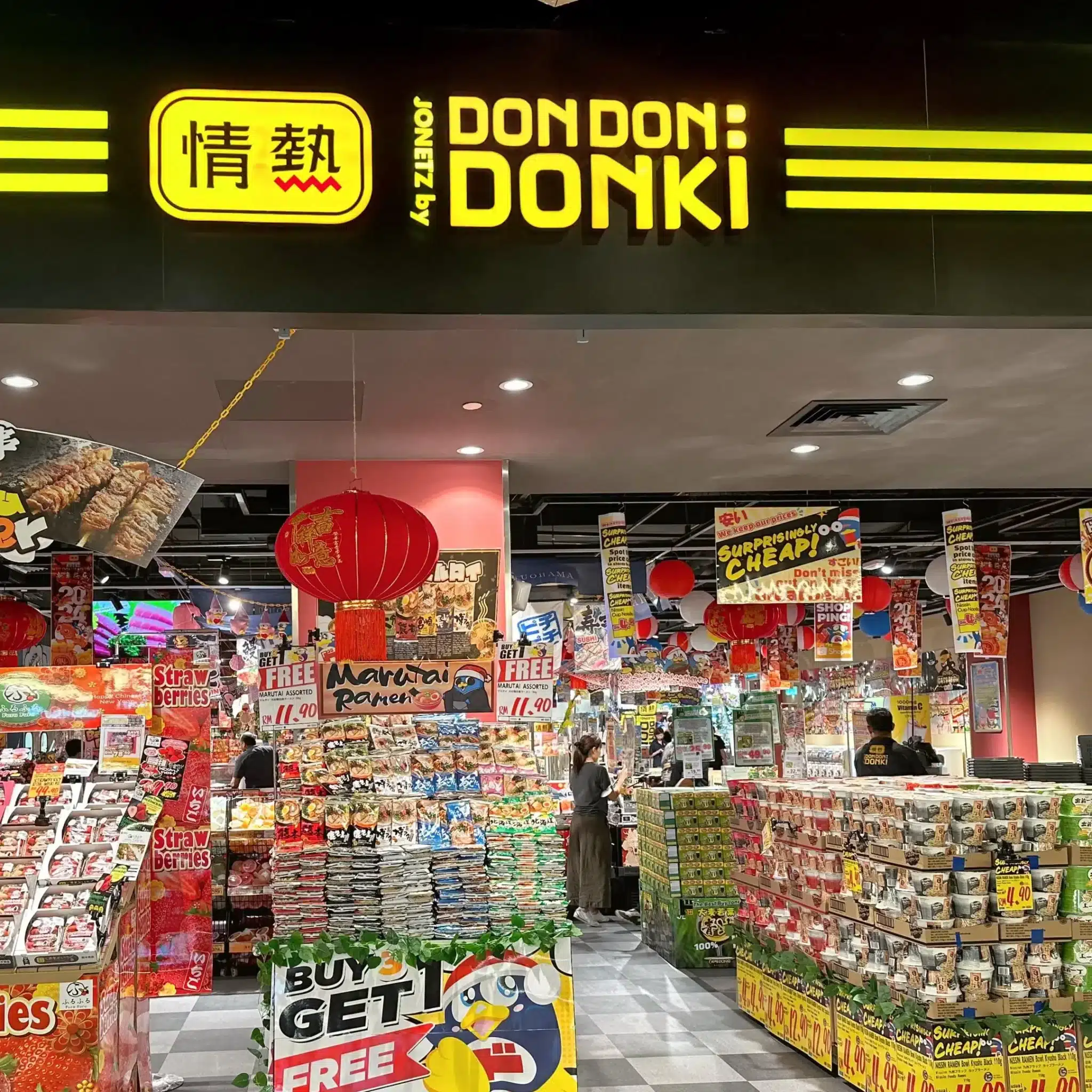 Colourful interior of a Don Don Donki store displays various packaged goods, snacks, and bottles of ramune, alongside promotional signs under bright lighting and red lanterns.