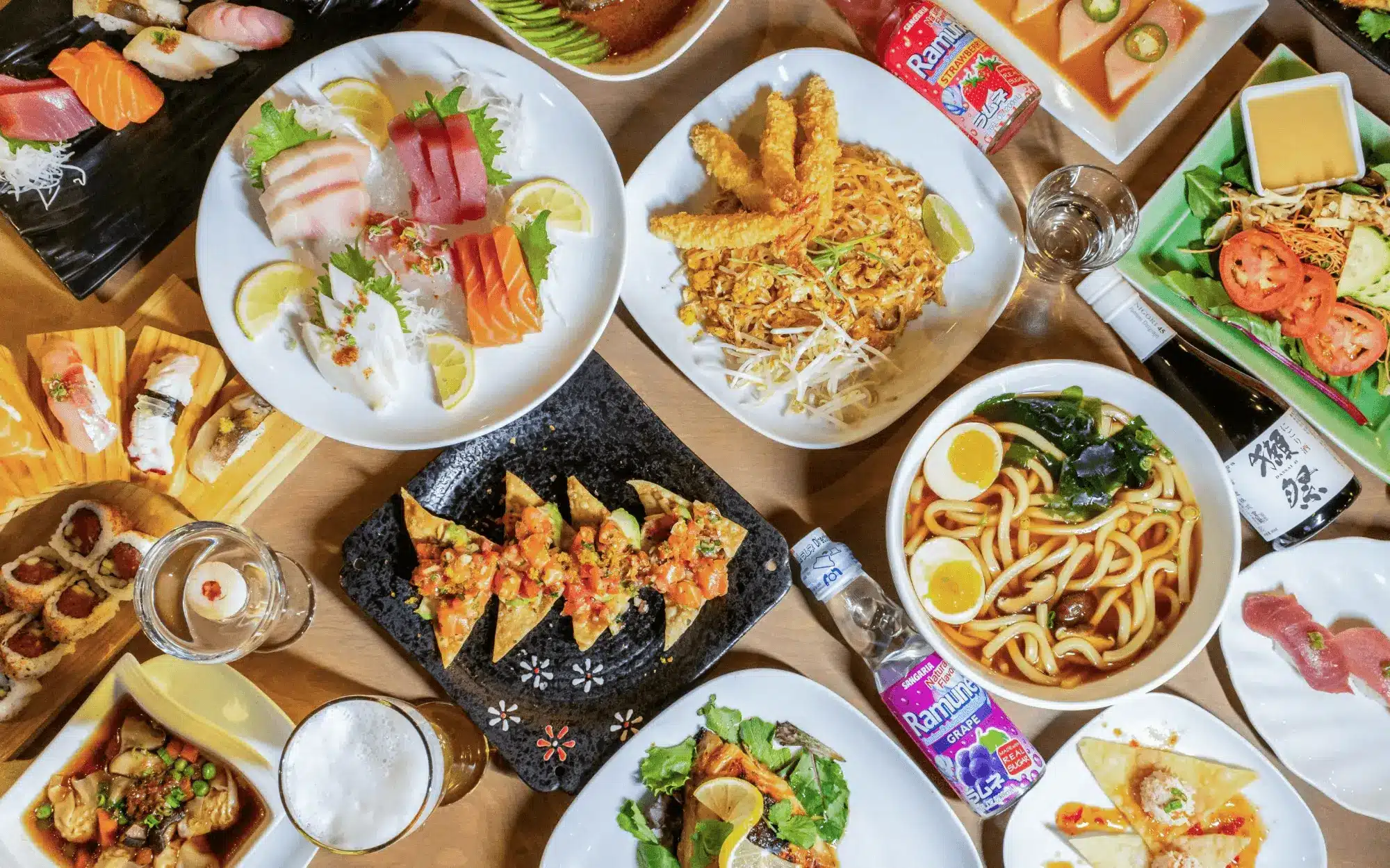 A variety of Japanese dishes, including sushi, sashimi, noodles, fried prawns, and refreshing ramune drinks, arranged on a wooden table viewed from above.