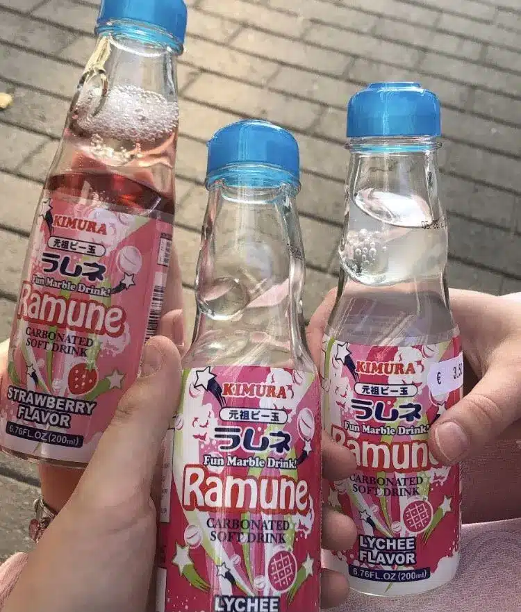 Three hands holding bottles of Kimura Ramune, the classic Japanese carbonated soft drink, in strawberry and lychee flavours. The bottles feature marble stoppers and pink labels, showcased on a paved outdoor surface.